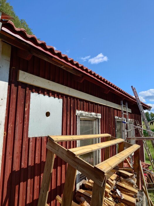 Röd stuga, byggnadsställning, träbitar, takpannor, fönster, klar himmel, under renovering eller konstruktion, lantlig omgivning.