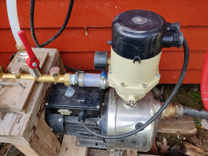Elmotor kopplad till pump med slangar, ventil och röda stugan, installation utomhus på träplattform.