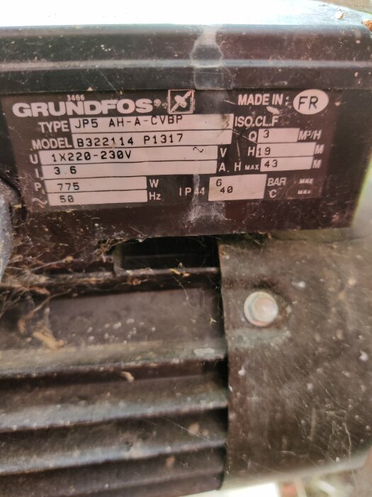 Sliten Grundfos pumpetikett, tekniska specifikationer, dammig yta med spindelnät. Made in France.