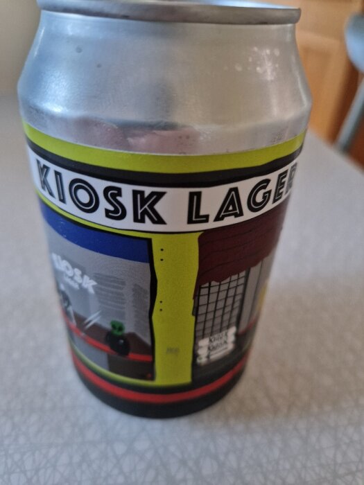 Burdruk på en ölburk med texten "KIOSK LAGER", färgglada streck och illustrationer, lutande perspektiv.