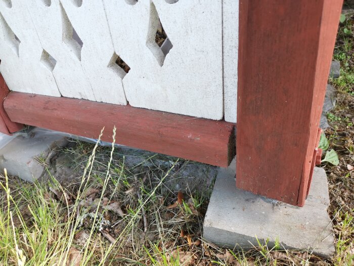 Vitt staket med utsågade mönster, röd brädbas, grå betongsten och grönt gräs.