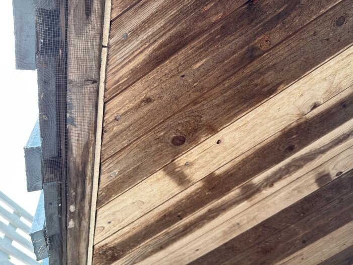 Trävägg och hörn av byggnad med nät och metall under solljus och skuggor.