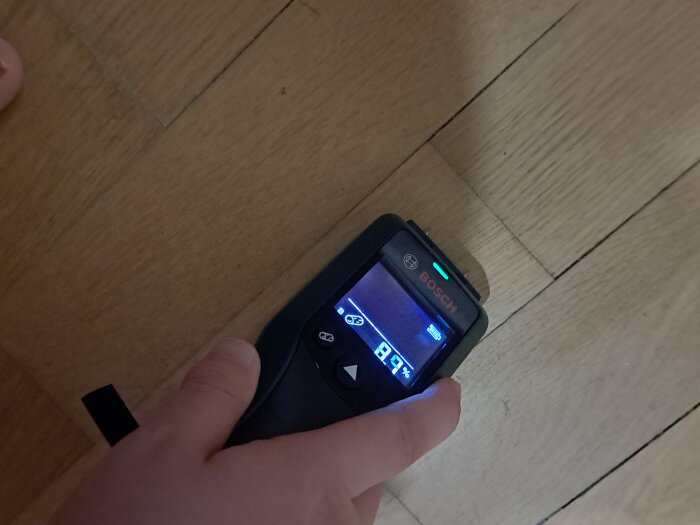 En hand håller en Bosch laseravståndsmätare mot ett trägolv; mäter, digital skärm synlig.