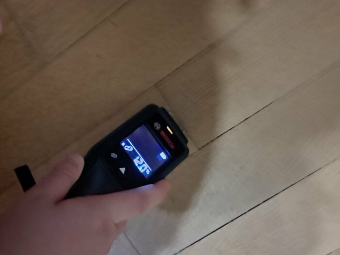En hand håller en infraröd termometer riktad mot trägolv; visar temperatur på display.