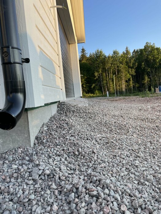 Byggnad med grå sidopanel och nedrör, grusgrund, skog i bakgrunden, blå himmel, dagsljus.