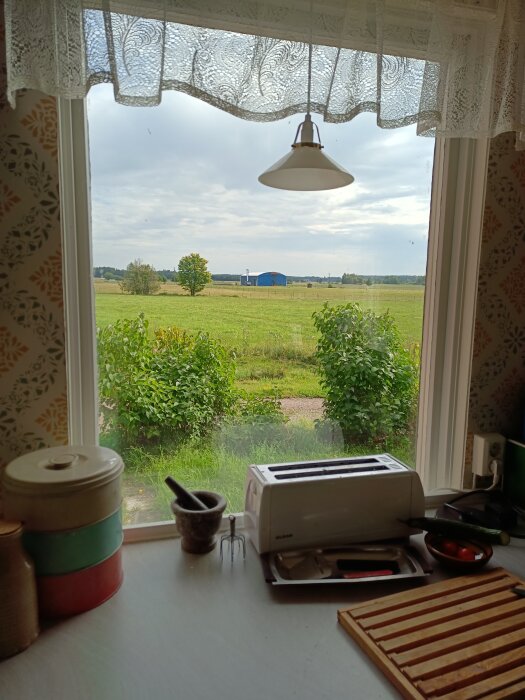 Kök med brödrost, mortel, burkar, sett genom fönstret mot landskap, lantlig vy, molnig himmel.
