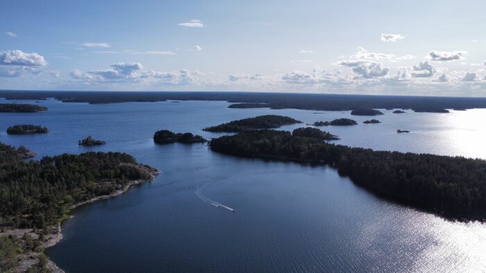 Flygbild över skärgård, solsken, båt skapar kölvatten, flera öar och blått vatten.