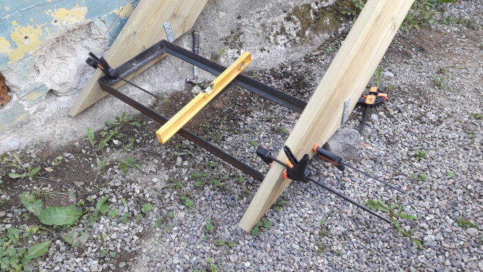 Träramkonstruktion fästs med skruvstäd och klämmor vid stenfylld mark och sliten blå vägg.