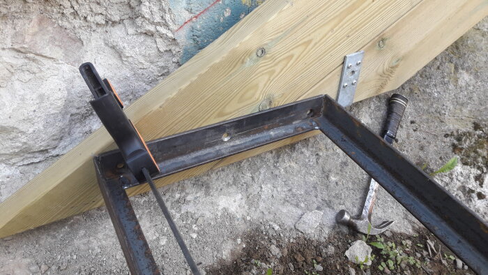 Träbjälke fäst mot betongvägg, klämmor, metallstång, metallbeslag, hammare, utomhus, byggnadsarbete, verktyg, konstruktion.