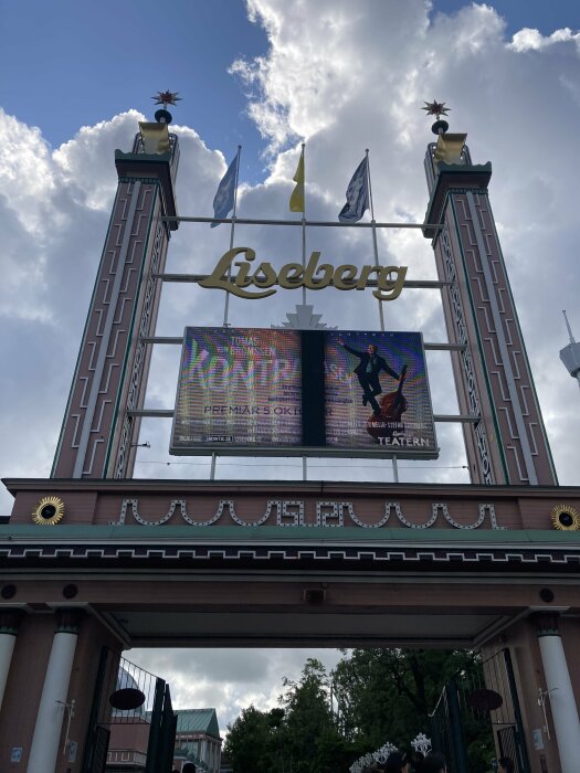 Entréskylt med ordet "Liseberg", flaggor, digital skärm, molnig himmel, främre arkitektoniska detaljer.