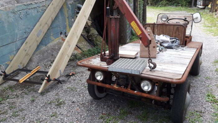 Gammalt fordon ombyggt till kranbil med trädetaljer, stålfälgar, klämma och betongplattor. Vintage, rostigt på grus.