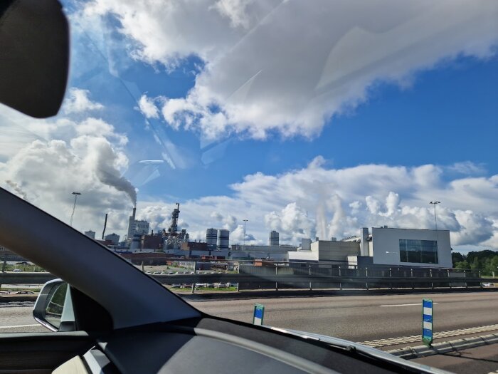 Industriområde med skorstenar, rök, fotograferat från bil, blå himmel och moln.