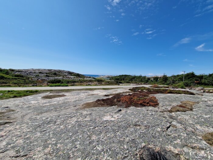 Klippig terräng, mossig vegetation, blå himmel, dagtid, utomhus, ingen synlig bebyggelse, väg i fjärran.