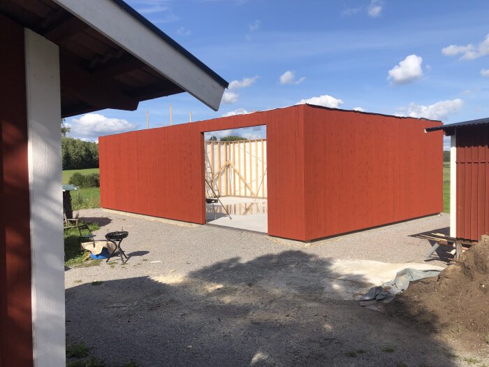 Röd träkonstruktion under byggnad, klar himmel, gårdsplan med grus, delar av byggnad i förgrunden.