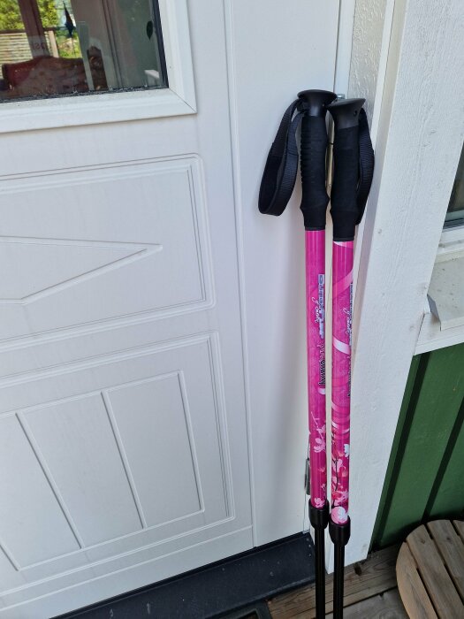 Två rosa vandringsstavar vid en vit dörr, handtag och remmar synliga, uteplatsmiljö.