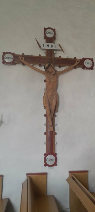 Ett träkors med en skulptur av Jesus, omgiven av symboler för evangelisterna, mot en ljus vägg.