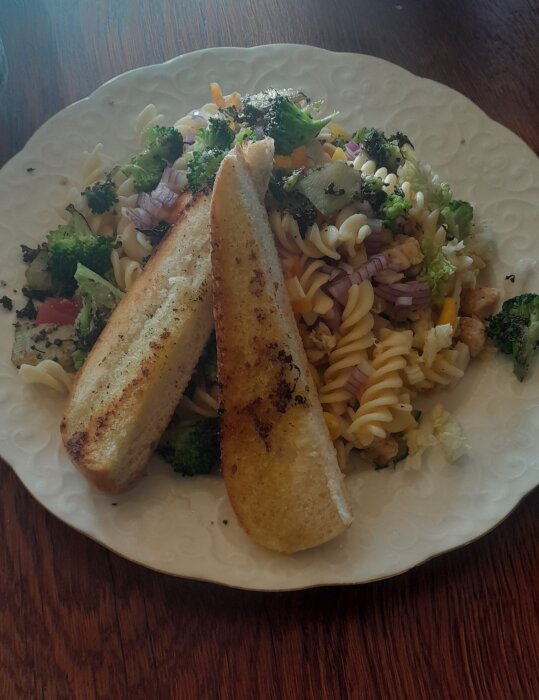 Pastasallad med broccoli, rödlök, vitlök och två skivor rostat bröd på träbord.