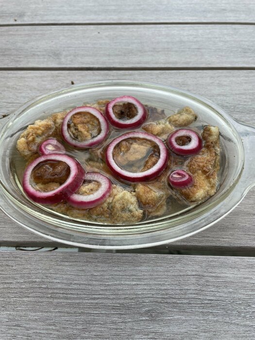 Stekt mat i klar glasskål toppad med rödlöksringar, på ett träbord utomhus.