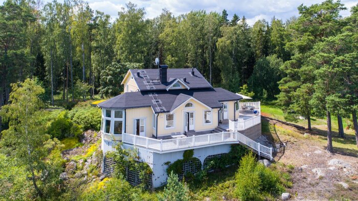 Gult hus med veranda omgivet av skog, solpaneler på taket, bilder taget från luften.