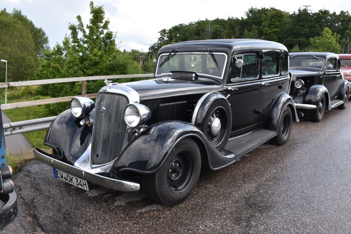 Klassiska bilar, svart, parkerade, vintage, kromdetaljer, registreringsskylt, träd i bakgrunden, dagtid, molnig himmel.