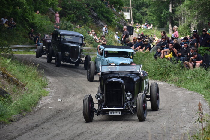 Gamla bilar kör på grusväg, åskådare tittar på längs sidorna, grönskande natur, sommardag, bilträff eller rally.