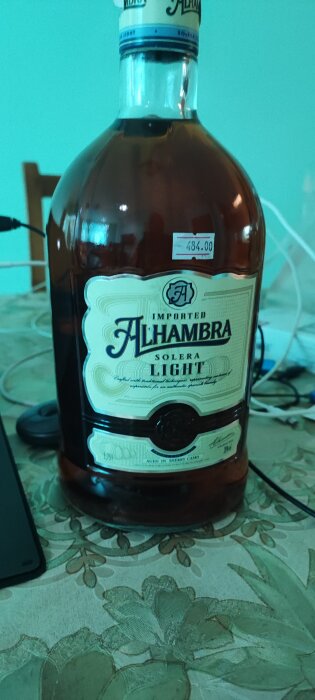 Flaska med mörkt innehåll, etikett "Alhambra Solera Light", prislapp på bord med mönstrad duk, bakgrundsföremål, smartphone.