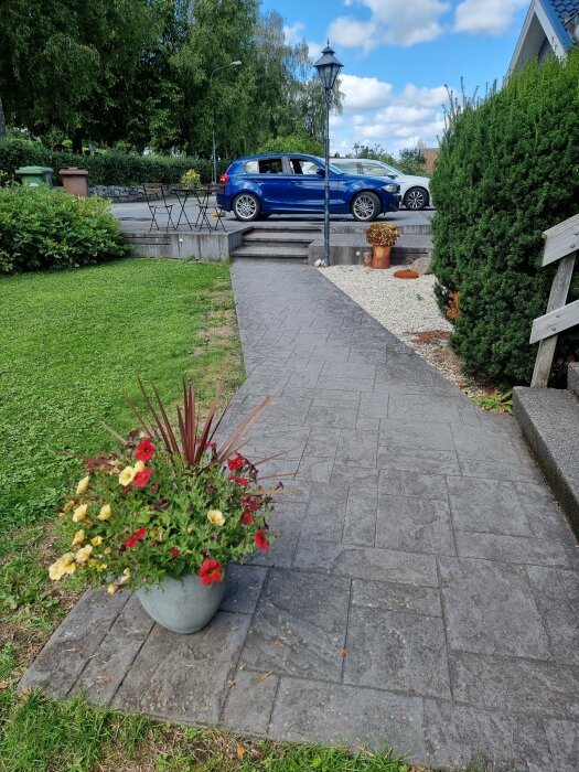 En stenlagd gångväg leder till en parkerad bil, bredvid gröna buskar och blommande krukväxter på en solig dag.