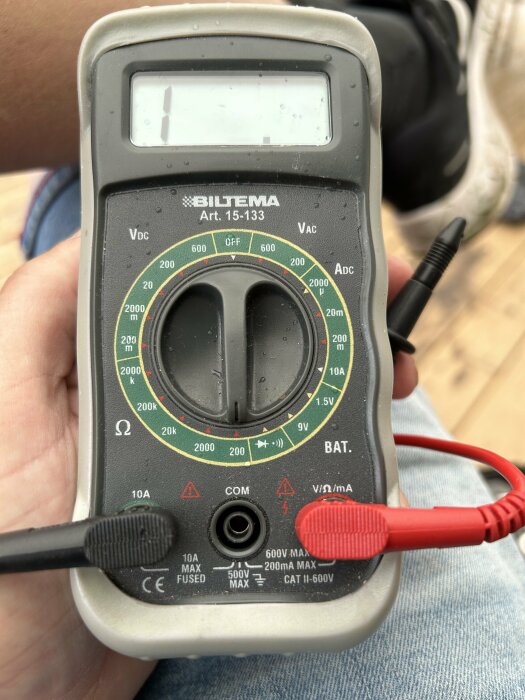 Digital multimeter hålls i hand, testledningar anslutna, skala för spänning och ström, mätverktyg, Biltema-märke.