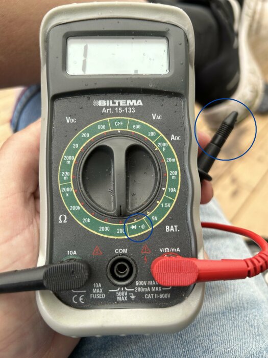 Multimeter hålls i hand, testsladdar anslutna, mätknapp inställd på batterispänningskontroll, otydlig display.