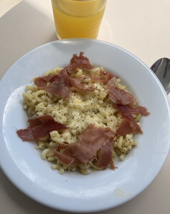 Pasta med ost och skinka, svartpeppar, juiceglas, på en vit tallrik, bestick, matbord.