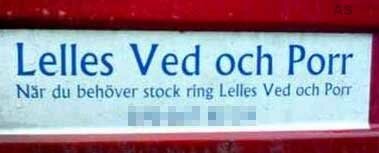 Skylt med text "Lelles Ved och Porr", röd bakgrund, delvis suddig, svensk humor, kombinerat erbjudande ved och vuxeninnehåll.