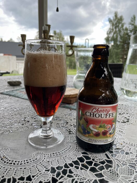 Ett glas med mörk öl och skum intill en tom ölflaska, på en spetsduk utomhus.