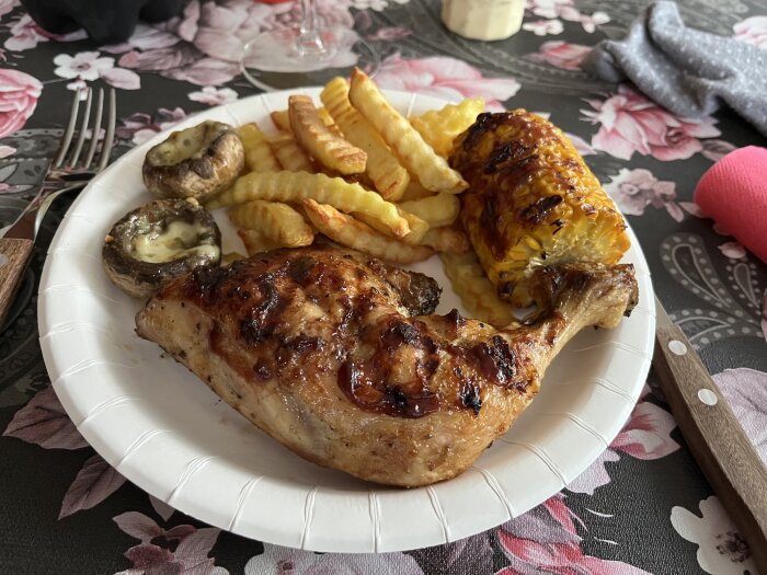 Grillad kycklinglår, pommes frites, majskolv, grillade champinjoner på en pappersservis med bestick vid sidan.