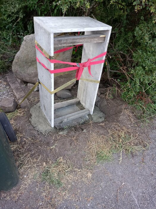 Avstängd skadad betongpapperskorg med varningsband, placerad ute vid en gångväg omgiven av vegetation.