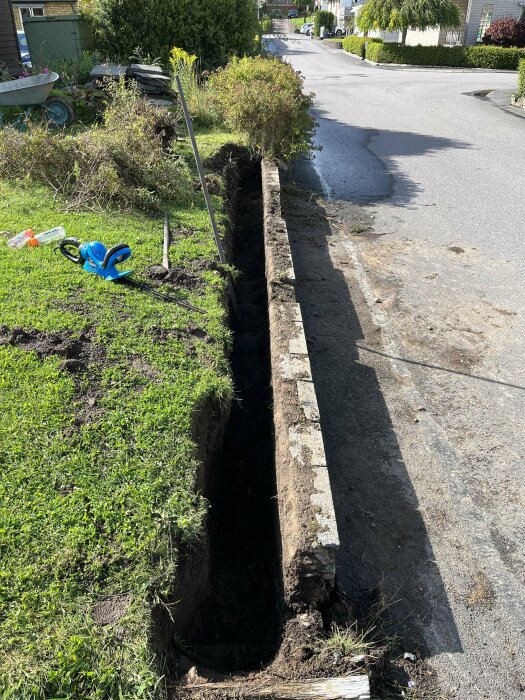 Lång grävd grävning vid gräsbevuxen kant, verktyg spridda, i bostadsområde med hus och vägar.