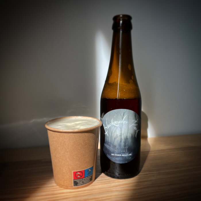 Ölflaska med stout, kaffemugg på träyta, dramatiskt ljus, skuggor, modern atmosfär.