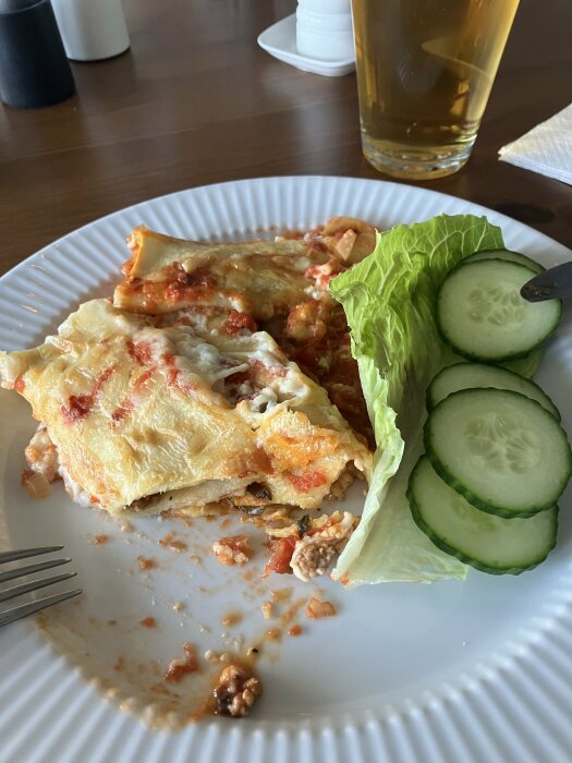 Halväten lasagne med isbergssallad och gurkskivor på vit tallrik, glas med dryck i bakgrunden.