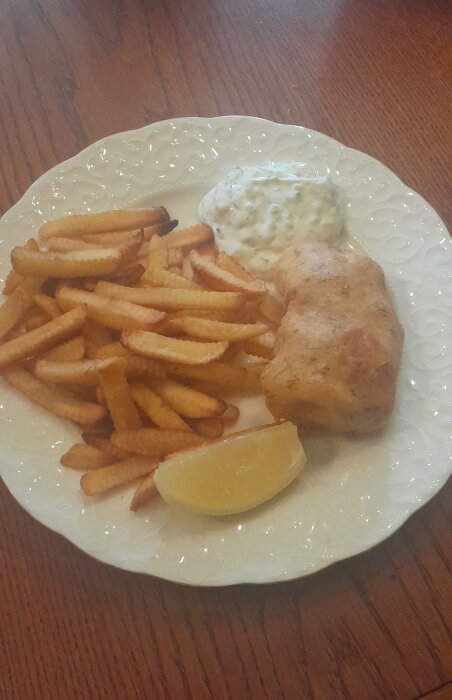 En tallrik med pommes frites, kycklingfilé, citronklyfta och remouladsås på ett träbord.