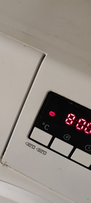 Digital termostat visar 21.5 grader Celsius på en vit vägg, med funktionssymboler och temperaturknappar.