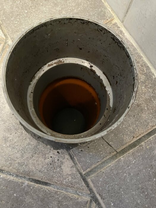 En smutsig, rostig metallhink med lite vätska på kakelgolv.