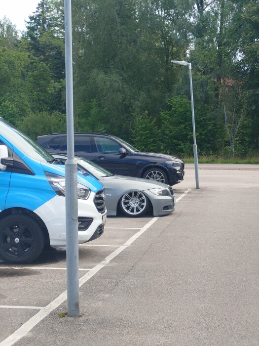 Parkering med bilar i rader, gatlyktor, gröna träd i bakgrunden, klar dag.