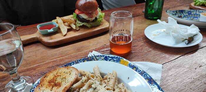 Måltid på träbord: burgare, pommes, pasta, öl, vattenglas, använd servett. Casual, avslappnad atmosfär.