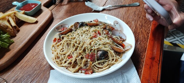 Spaghetti med skaldjur, riven ost, bladpersilja på träunderlägg, pommes och ketchup i bakgrunden, person håller mobiltelefon.