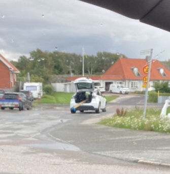 Suddig bild. Kompakt elfordon. Parkeringsplats. Röda byggnader. Skylt med bilsymbol. Gråa moln. Fläckar som antyder regnstänk på en ruta.