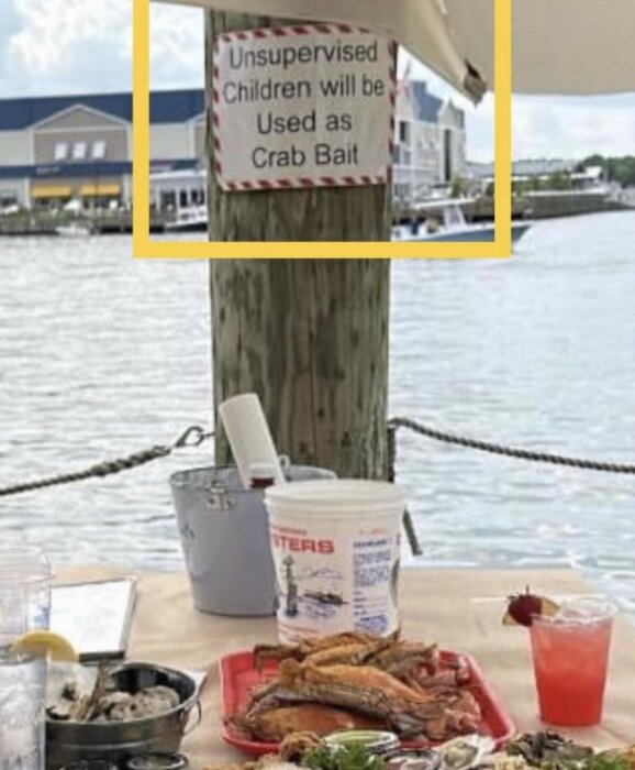 Skylt varnar "Unsupervised Children will be Used as Crab Bait", mat på bord vid vattnet.
