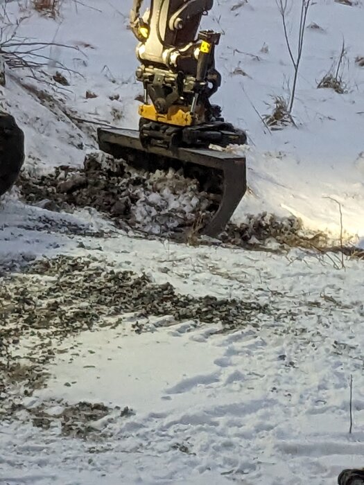 Grävmaskin med band rör sig i snötäckt terräng, uppgrävd mark och stenar, solnedgång eller soluppgång i bakgrunden.