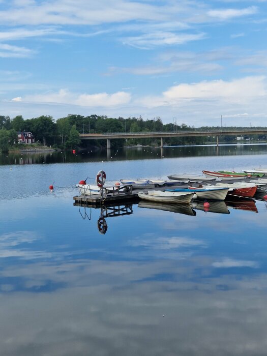 Brygga med roddbåtar, spegelblank sjö, bro i bakgrunden, moln speglas i vattnet, lugn stämning.