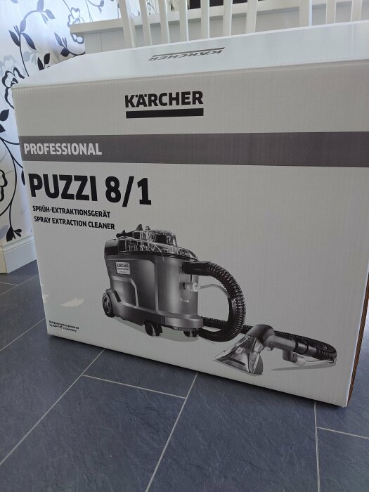 Kärcher Professional Puzzi 8/1-rengöringsmaskinsförpackning på golv framför vit staket.