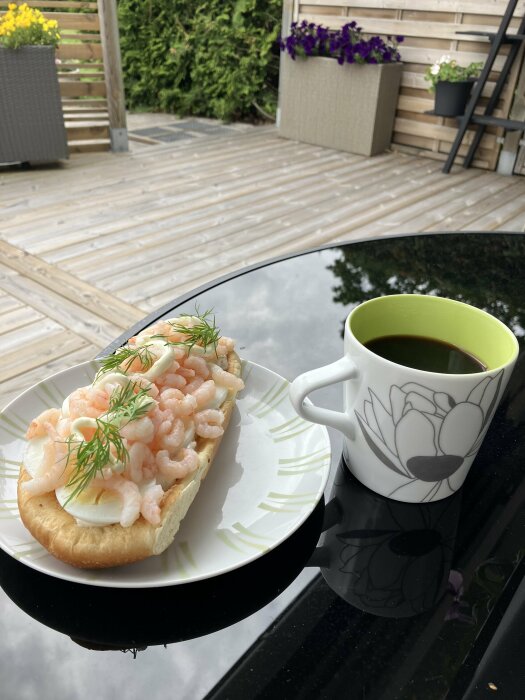 Fika utomhus med räkmacka och kaffe, träterrass, blommor i bakgrunden, reflekterande bordsover yta.