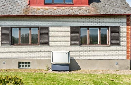 Ett hus med tegeltak, tegelstenar, vita fönster, en värmepump framför på gräsmattan.